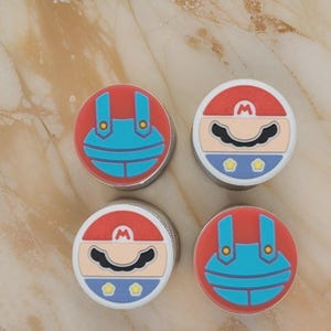 May include: Four circular metal containers with cartoon character designs. Two feature a red background with blue overalls, and two feature a red hat, black moustache, and blue overalls. The containers have a silver metal rim.