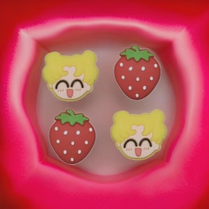 May include: Four colourful charms: two strawberry designs and two with a cartoon character face. The strawberries are red with green tops and white dots. The cartoon faces are yellow with a smiling expression. The charms are set in a pink box.