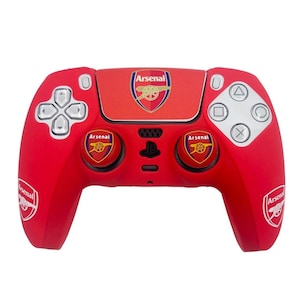 May include: Red video game controller featuring the Arsenal logo. The controller has silver buttons and a directional pad. The Arsenal logo is displayed on the top center and on the two thumbstick buttons. The Arsenal logo is also on the left and right sides of the controller.