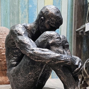 Puede incluir: Escultura de color bronce de una figura sentada, con las rodillas levantadas y la cabeza inclinada. La estatua tiene una superficie texturizada y una pose contemplativa. El fondo presenta una pared de madera con rayas verticales azules, verdes y blancas.