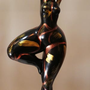 Può includere: Scultura nera di una persona in posa yoga, con le braccia sollevate sopra la testa e una gamba piegata. La scultura ha accenti dorati e rossi e poggia su una base nera.
