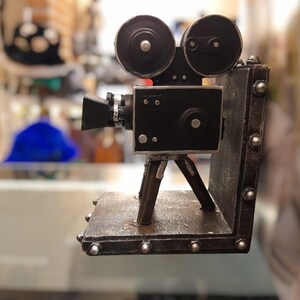 Puede incluir: Una cámara de cine negra de estilo vintage, con dos componentes en forma de carrete en la parte superior, montada sobre un trípode. La cámara está sobre una base rectangular con tachuelas metálicas decorativas. El diseño evoca una estética clásica y antigua.