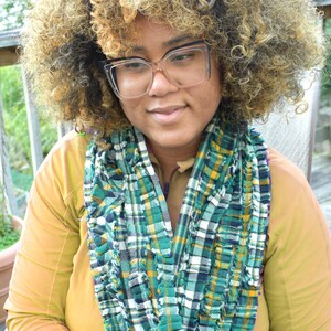 May include: A person wearing a green, white, and yellow plaid scarf. The scarf is draped over a mustard-colored long-sleeved shirt. The person is wearing clear-framed glasses and has curly, light brown hair.
