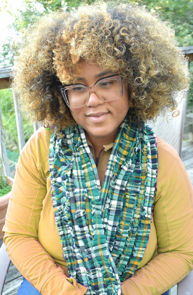 May include: A person wearing a green, white, and yellow plaid scarf. The scarf is draped over a mustard-colored long-sleeved shirt. The person is wearing clear-framed glasses and has curly, light brown hair.