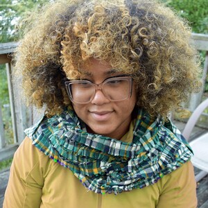 May include: A green, blue, and white plaid infinity scarf. The scarf is made of a soft fabric and is wrapped around the neck. The person is wearing a yellow jacket and clear-framed glasses.