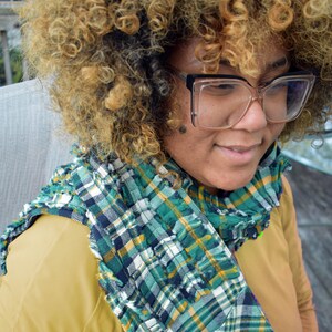 May include: A green, white, and navy blue plaid scarf with a ruffled edge. The scarf is draped over a mustard-colored top. The person in the image is wearing large, clear-framed glasses.
