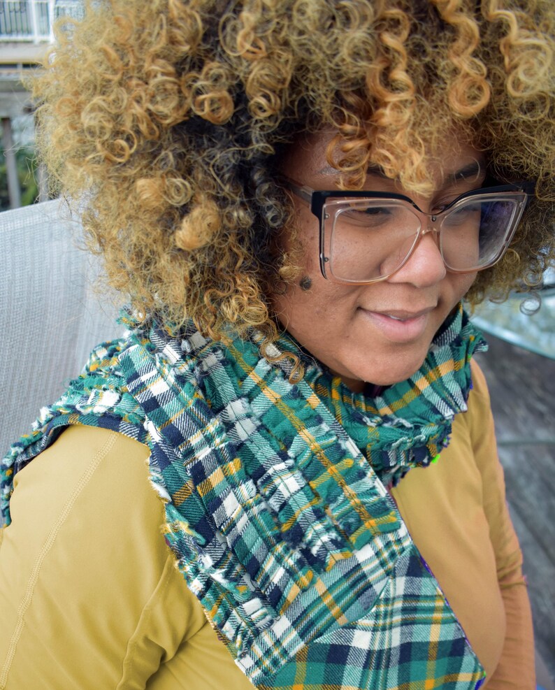 May include: A green, white, and navy blue plaid scarf with a ruffled edge. The scarf is draped over a mustard-colored top. The person in the image is wearing large, clear-framed glasses.