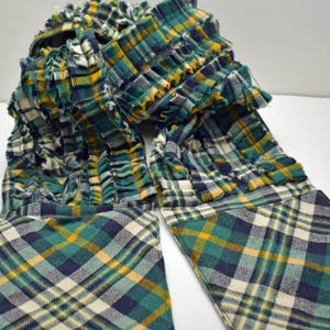 May include: A green, yellow, and navy blue plaid scarf with a ruffled design. The scarf is made of a soft fabric and has a unique, textured appearance. The plaid pattern is visible throughout the scarf.