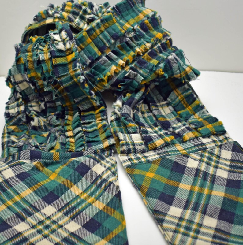 May include: A green, yellow, and navy blue plaid scarf with a ruffled design. The scarf is made of a soft fabric and has a unique, textured appearance. The plaid pattern is visible throughout the scarf.