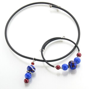 Può includere: Un set di due collane choker nere con un ciondolo con perline di vetro blu e rosse. Il ciondolo è attaccato alla collana più lunga.