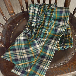May include: A green, navy, yellow, and white plaid scarf with a ruffled edge is draped over a wooden chair. The scarf is made of a soft fabric and features a classic plaid pattern. The chair has a dark brown wooden frame and a leather seat.