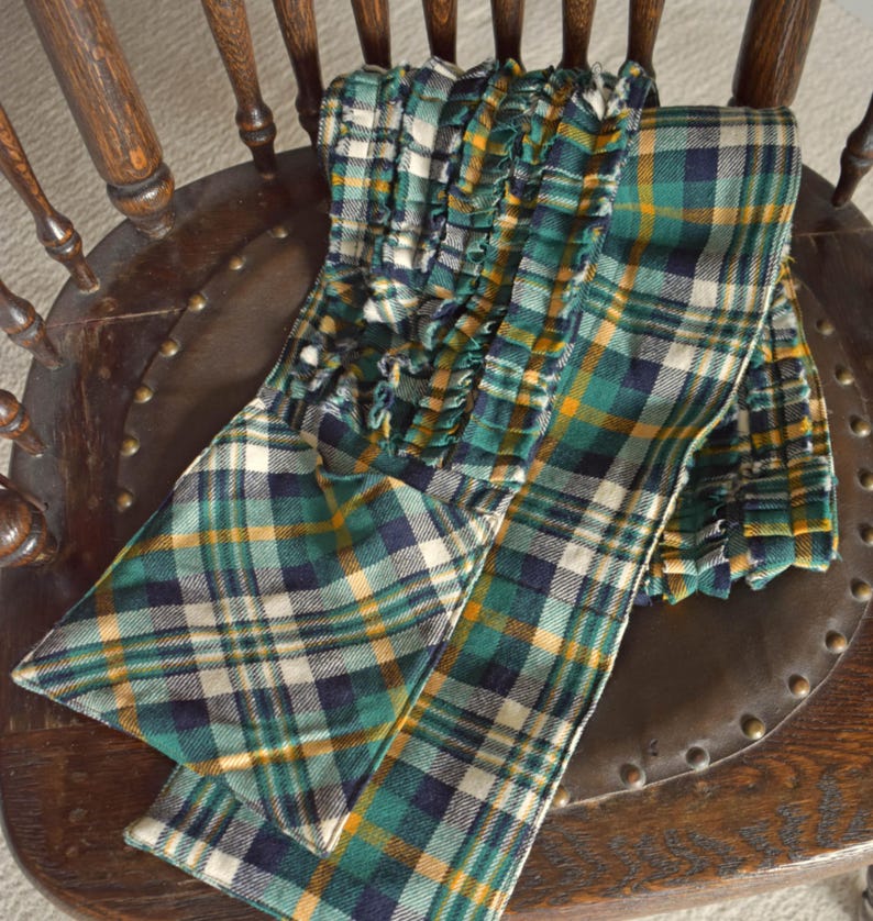 May include: A green, navy, yellow, and white plaid scarf with a ruffled edge is draped over a wooden chair. The scarf is made of a soft fabric and features a classic plaid pattern. The chair has a dark brown wooden frame and a leather seat.
