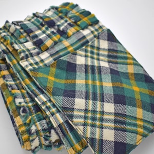 May include: A stack of folded plaid fabric items in shades of green, yellow, navy, and white. The fabric has a textured appearance and a classic plaid pattern. The edges of the fabric have a fringed detail.