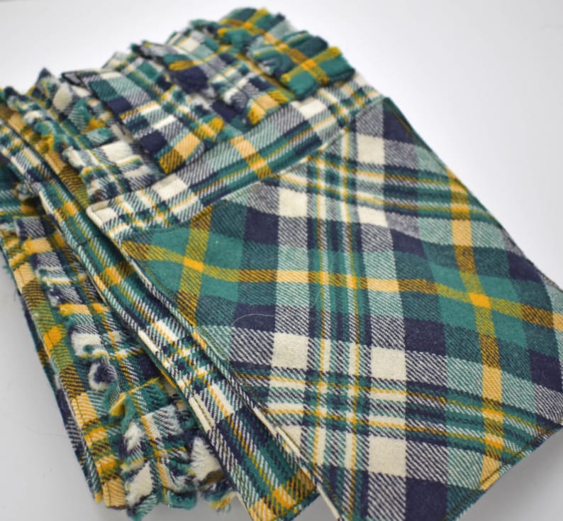 May include: A stack of folded plaid fabric items in shades of green, yellow, navy, and white. The fabric has a textured appearance and a classic plaid pattern. The edges of the fabric have a fringed detail.