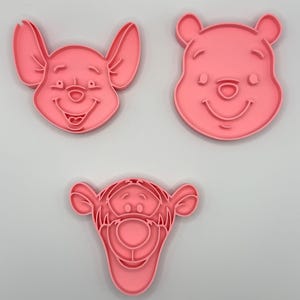 Pode incluir: Tr&ecirc;s cortadores de biscoitos de pl&aacute;stico rosa com os rostos de Leit&atilde;o, Ursinho Pooh e Tigr&atilde;o. Os cortadores t&ecirc;m a forma dos rostos dos personagens, com detalhes. Ideais para fazer biscoitos tem&aacute;ticos.