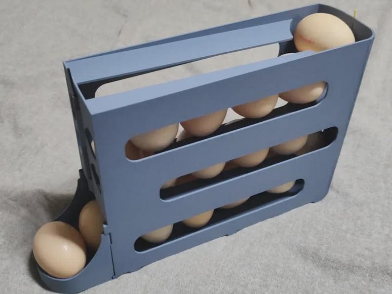 May include: A blue plastic egg holder with multiple slots, filled with beige eggs. The holder is designed to store and dispense eggs, keeping them organized. The design allows for easy access and visibility of the eggs.