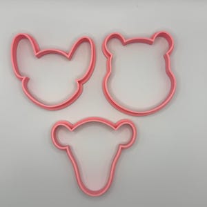 Pode incluir: Tr&ecirc;s cortadores de biscoitos de pl&aacute;stico rosa em forma de rostos de animais. As formas incluem um rato, um urso e um veado. Os cortadores de biscoitos est&atilde;o em um fundo branco.