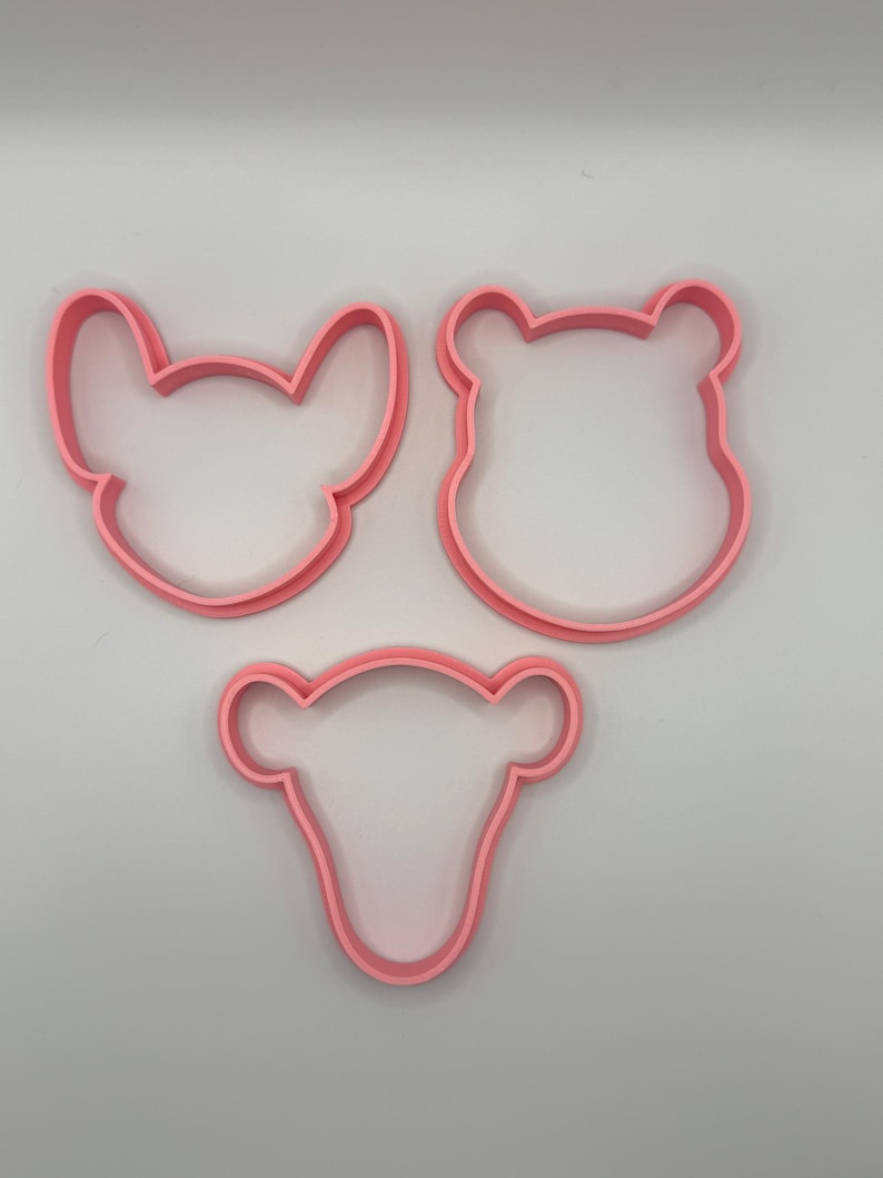 Pode incluir: Tr&ecirc;s cortadores de biscoitos de pl&aacute;stico rosa em forma de rostos de animais. As formas incluem um rato, um urso e um veado. Os cortadores de biscoitos est&atilde;o em um fundo branco.