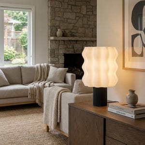 May include: A living room scene featuring a modern lamp with a white, wavy shade and a black base. The lamp sits on a dark wood cabinet, next to books and a small vase. A beige sofa with throw pillows and a blanket is in the background.