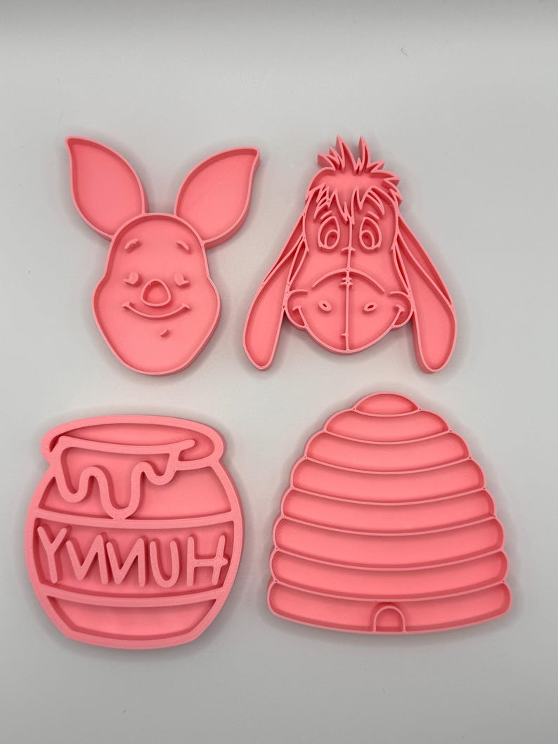Pode incluir: Quatro cortadores de biscoitos cor-de-rosa com personagens e s&iacute;mbolos de uma hist&oacute;ria infantil popular. O conjunto inclui uma cara de coelho, uma cara de burro, um pote de mel com a palavra "YMMUH" e uma colmeia.