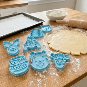 Pode incluir: Cortadores de biscoitos azuis claros em v&aacute;rias formas, incluindo personagens de Winnie the Pooh e um pote de mel. Est&atilde;o dispostos numa superf&iacute;cie de madeira com massa estendida, farinha e um rolo. Uma assadeira est&aacute; no fundo.