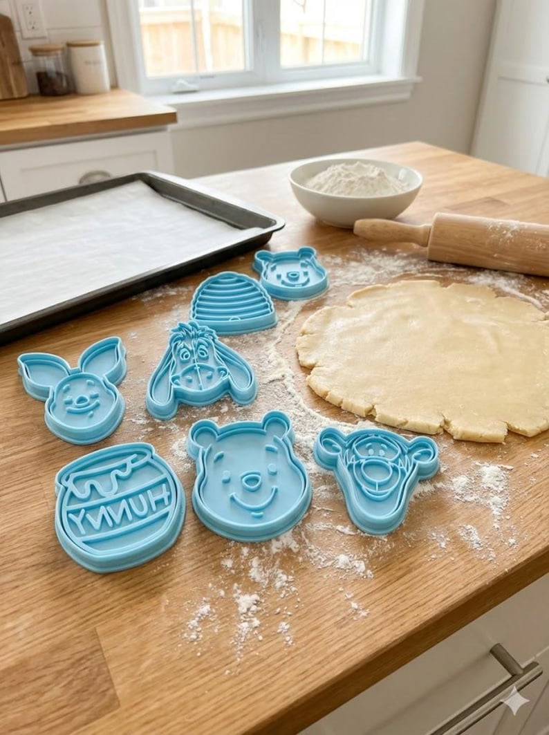 Pode incluir: Cortadores de biscoitos azuis claros em v&aacute;rias formas, incluindo personagens de Winnie the Pooh e um pote de mel. Est&atilde;o dispostos numa superf&iacute;cie de madeira com massa estendida, farinha e um rolo. Uma assadeira est&aacute; no fundo.