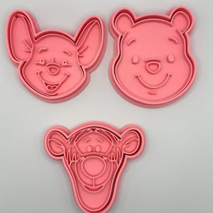 Pode incluir: Tr&ecirc;s cortadores de biscoitos de pl&aacute;stico rosa com rostos de personagens de desenhos animados. O conjunto inclui um urso sorridente, um porco sorridente e um tigre sorridente. Os cortadores de biscoitos s&atilde;o projetados com tra&ccedil;os faciais detalhados.