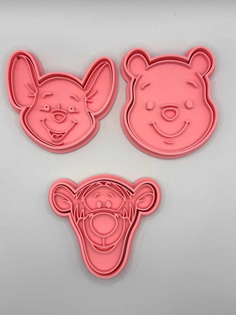 Pode incluir: Tr&ecirc;s cortadores de biscoitos de pl&aacute;stico rosa com rostos de personagens de desenhos animados. O conjunto inclui um urso sorridente, um porco sorridente e um tigre sorridente. Os cortadores de biscoitos s&atilde;o projetados com tra&ccedil;os faciais detalhados.