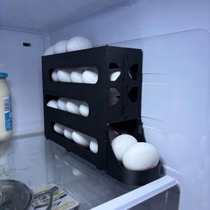 May include: A black, three-tiered egg holder filled with white eggs inside a refrigerator. The egg holder is made of plastic and has a sliding drawer at the bottom. The refrigerator is white and has a light inside.