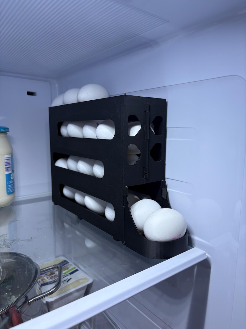 May include: A black, three-tiered egg holder filled with white eggs inside a refrigerator. The egg holder is made of plastic and has a sliding drawer at the bottom. The refrigerator is white and has a light inside.
