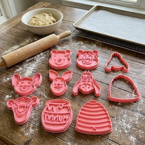 Pode incluir: Cortadores de biscoitos cor-de-rosa em v&aacute;rias formas, incluindo personagens de Winnie the Pooh, um pote de mel e uma colmeia, dispostos sobre uma superf&iacute;cie de madeira polvilhada com farinha. Um rolo e uma tigela com massa tamb&eacute;m est&atilde;o presentes, sugerindo assar.