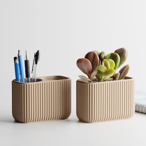 May include: Two beige, ribbed desk organizers. One holds pens and pencils, the other a small succulent plant. The organizers are rectangular with rounded corners, and sit on a white surface.