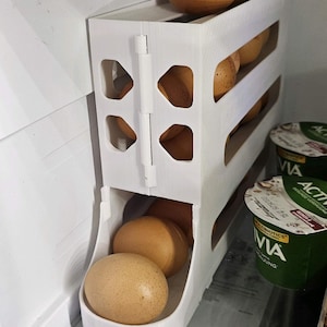 May include: A white, 3-tier egg holder inside a refrigerator. The holder is filled with brown eggs. The design features a hinged door and open sides for easy access. The refrigerator also contains yogurt containers.
