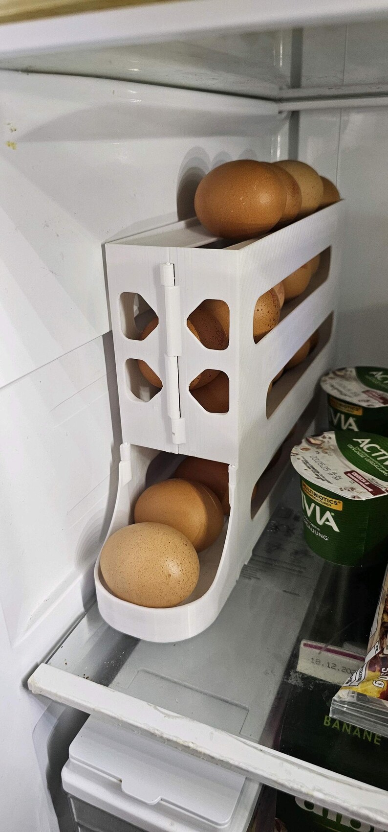 May include: A white, 3-tier egg holder inside a refrigerator. The holder is filled with brown eggs. The design features a hinged door and open sides for easy access. The refrigerator also contains yogurt containers.