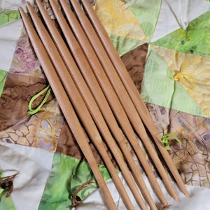 May include: A collection of eight wooden knitting needles, each with a tapered point. The needles are arranged on a colorful patchwork quilt with green, yellow, and brown patterns. The needles are made of light-colored wood.