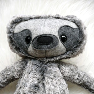 May include: A gray plush sloth toy with black eyes and a black nose. The toy is made of soft, fuzzy fabric and has a friendly expression.