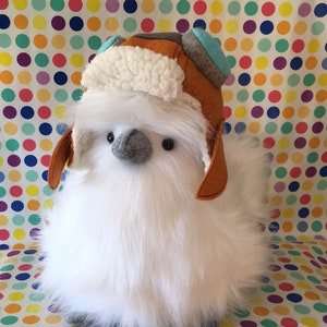 Może przedstawiać: Biała pluszowa zabawka z brązową i niebieską czapką lotniczą z nausznikami. Zabawka ma szary dziób i czarne oczy.