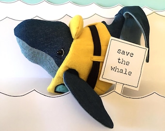 May include: A blue whale plush toy wearing a yellow life vest and holding a sign that says "save the whale". The whale is floating in a white sea with three white birds flying above.