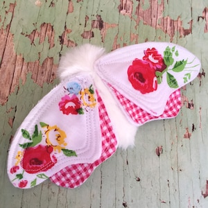 Puede incluir: Una mariposa de tela blanca con ribete de vichy rosa y blanco y alas de tela floral. La mariposa tiene un cuerpo blanco esponjoso.
