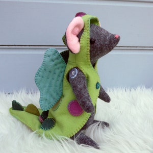 May include: A green felt mouse wearing a green felt dragon costume with pink felt ears and a blue felt wing. The mouse is sitting on a white fluffy surface.