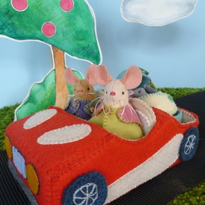 May include: Three felt mice in a red toy car with white trim. The car has a white steering wheel and black wheels. The mice are wearing colorful clothes and have embroidered details.