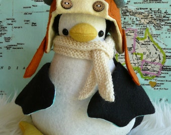 May include: A plush penguin wearing a brown, orange, and blue aviator hat with a white knit scarf. The penguin is sitting on a white fluffy surface.