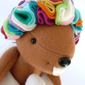 May include: A brown felt toy beaver wearing a colourful felt flower crown. The crown is made of multiple layers of felt in various colours, including pink, green, blue, yellow and orange.