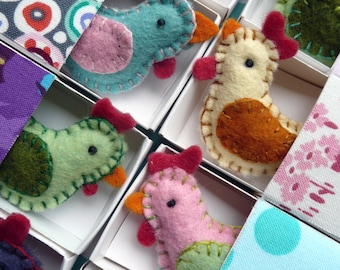 May include: Handmade felt bird ornaments in various colors, including blue, pink, green, and cream. Each bird has a contrasting wing and beak, with red accents. The birds are displayed in individual white boxes, with patterned fabric dividers.