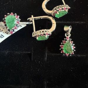 May include: A jewellery set comprising a ring, earrings, and a pendant. The items have a silver finish and are decorated with green emeralds and small red and blue gemstones. The earrings have a lever back closure.