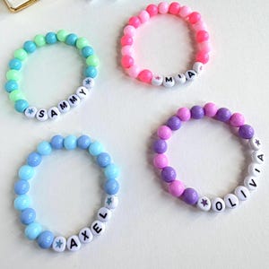 May include: Four beaded bracelets in shades of blue, green, pink, and purple. Each bracelet features white letter beads spelling out a name: Sammy, Mia, Axel, and Olivia. The bracelets are made of small, round beads.