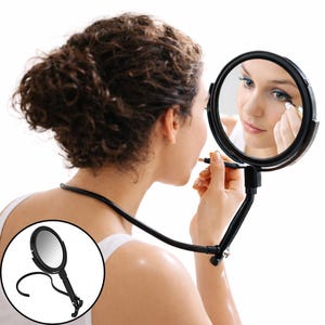 May include: A black, flexible, hands-free makeup mirror with a round mirror and a clip-on base. The mirror is being used by a person applying makeup. The mirror is designed to be worn around the neck for easy use.