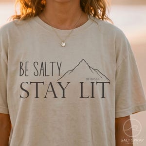 以下が含まれることがあります： 「BE SALTY STAY LIT」という黒い文字、山の線画、そして小さな「MATTHEW 5:13-14」という文字がプリントされたベージュの半袖Tシャツ。クルーネックでゆったりとしたフィット感のデザインです。