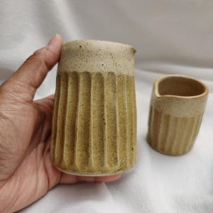 May include: Two handmade ceramic creamers. The larger creamer has a ribbed design on the lower portion and a speckled, light-colored rim. The smaller creamer has a similar design. Both are in a neutral, earthy tone.