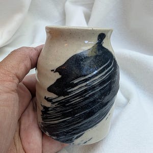 May include: Handmade ceramic vase with a beige base and a bold black brushstroke design. The small, cylindrical vase has a slightly flared rim and a textured surface, showcasing a unique artistic style. Perfect for displaying small flowers or as a decorative accent.